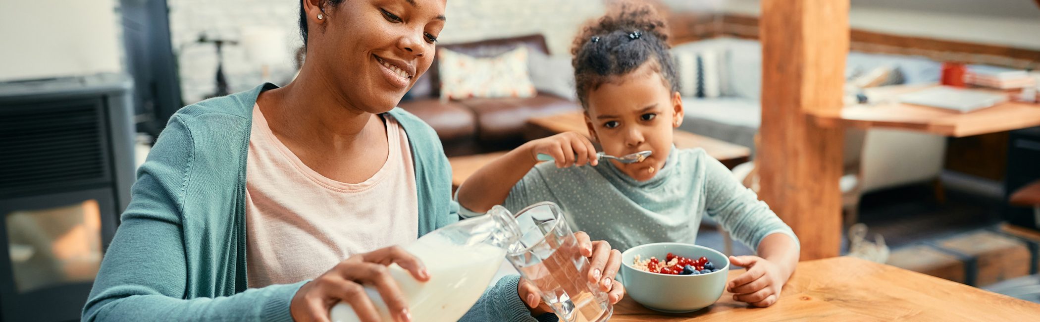 Happy African American mother pouring milk into a glass while eating breakfast with her daughter at dining table.